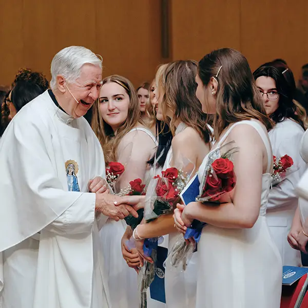 Girls at graduation wearing white dresses holding red roses shaking the priest's hand smiling.