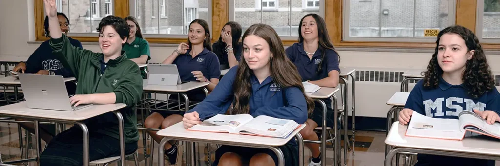 A group of girls sitting at their desks in a classroom. One girl has he hand raised.