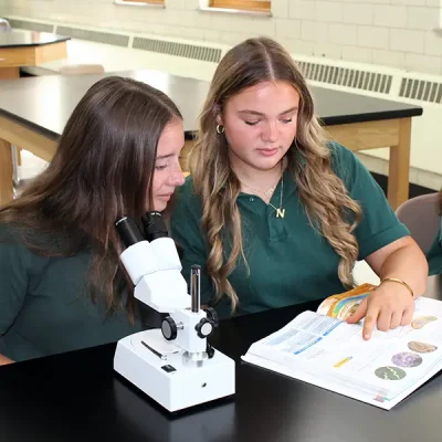 two girls in uniform sitting at a lab table looking at a textbook