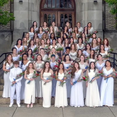 Photo of graduating class of MSM students standing in front of the school building wearing white dresses holding red roses