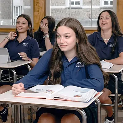 A group of girls sitting at their desks in a classroom. One girl has he hand raised.