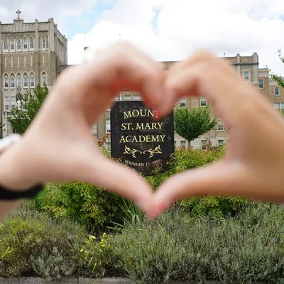 A pair of hands making a heart shape around the Mount St. Mary Academy sign in front of the school