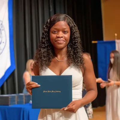 Graduate standing in the MSM auditorium holding her diploma in a white dress